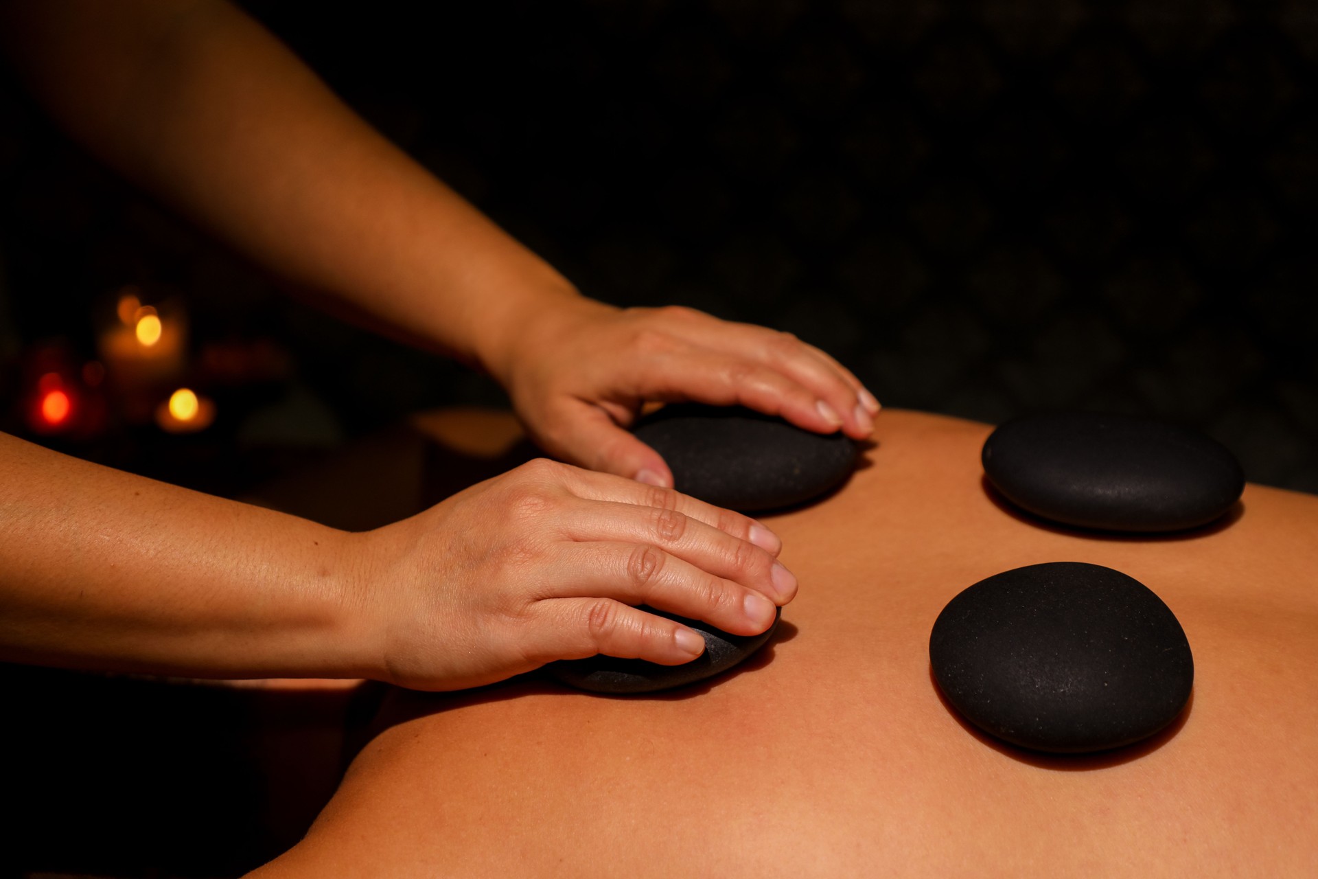 Woman's hands giving man stone therapy using black hot stones in Thai massage parlor by candlelight